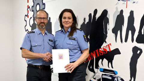 Neue Schutzfrau vor Ort bei der Polizeistation Melsungen; Ein Mann überreicht einer Frau eine Urkunde mit Hessewappen darauf, beide stehen vor einer weißen Wand mit zahlreichen Silhouetten