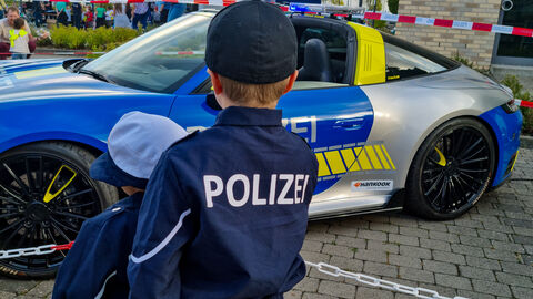 Kleiner Kinderkommissar in Polizeikostüm von hinten Fotografiert steht vor Polizeiporsche
