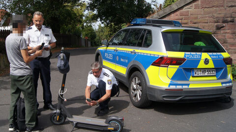 Zwei Polizeibeamte kontrollieren einen Mann mit seinem E-Scooter. Im Hintergrund steht ein Streifenwagen 