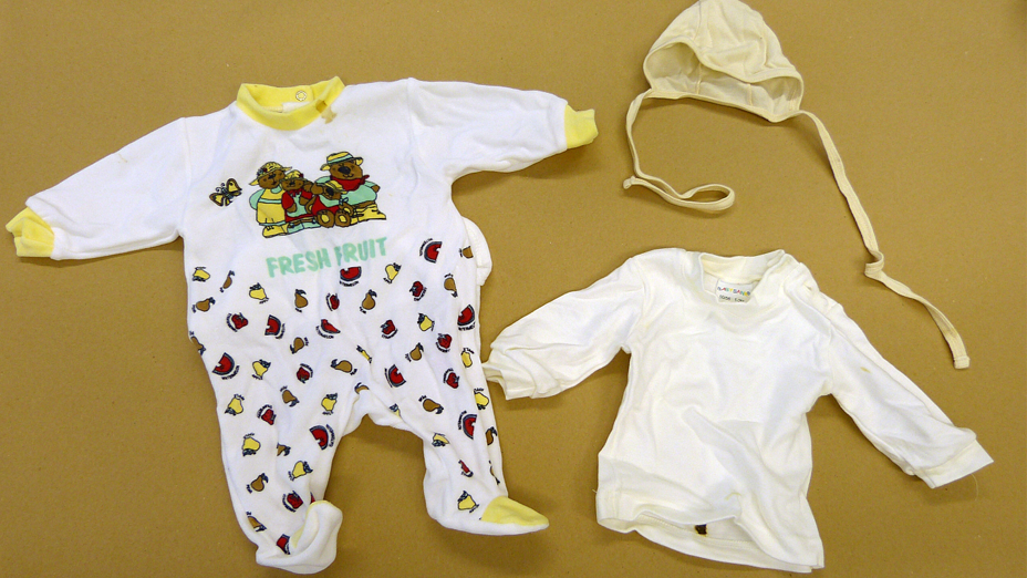 Links im Bild ein Babystrampler in der Grundfarbe weiß mit gelben Ärmelbündchen und Halseinfassung, in der oberen Hälfte des Stramplers ein Bild mit drei Teddybären und ein Schmetterling, in der unteren Hälfte des Stramplers mehrere  kleine Fruchtstücke in rot, braun und gelb, unter dem Bild der Schriftzug FRESH FRUIT, rechts eine hellgelbe Babymütze und ein Baby-Langarm-T-Shirt